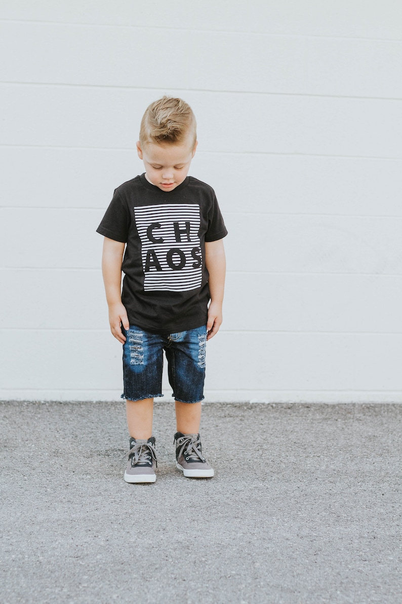 May include: A young boy wearing a black t-shirt with white stripes and the word "CHAOS" printed on it. He is also wearing blue denim shorts with distressed details and gray high-top sneakers.