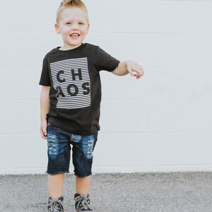 May include: A young boy wearing a black t-shirt with the word "CHAOS" printed in white on it. He is also wearing blue denim shorts and gray and black high-top sneakers.