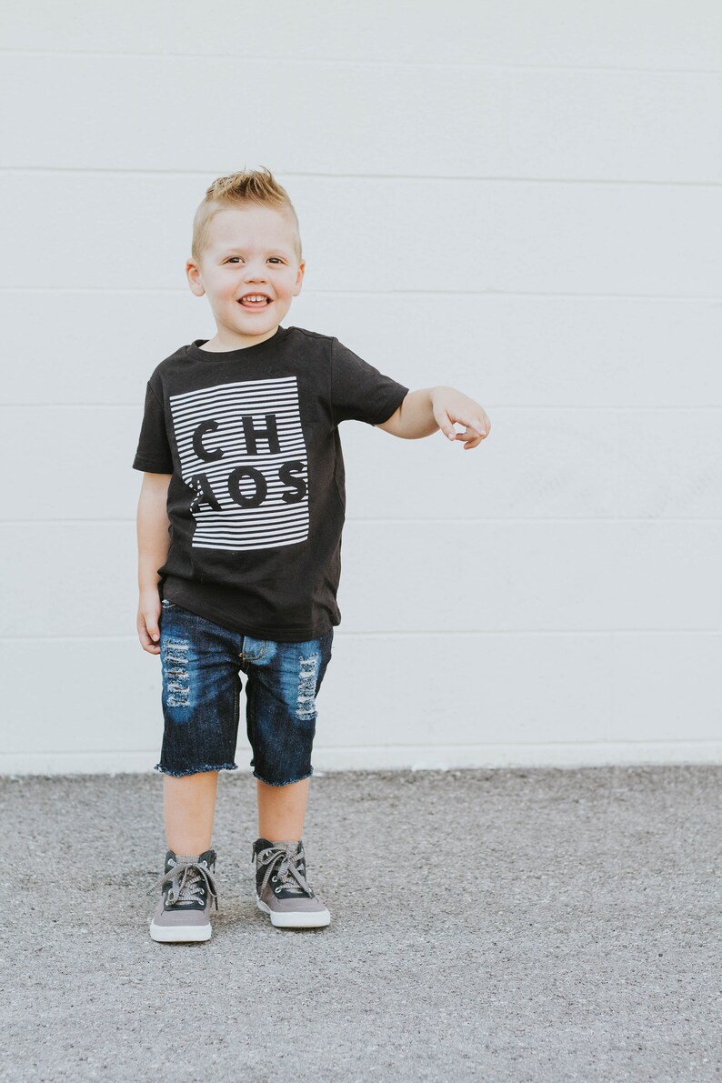 May include: A young boy wearing a black t-shirt with the word "CHAOS" printed in white on it. He is also wearing blue denim shorts and gray and black high-top sneakers.