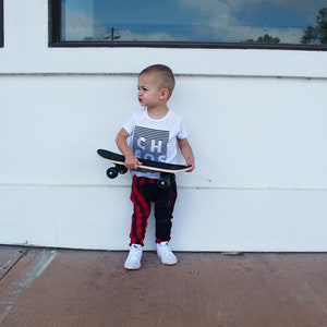 May include: A young boy wearing a white t-shirt with the word "Chaos" printed on it, black and red plaid pants, and white sneakers, holds a wooden skateboard.