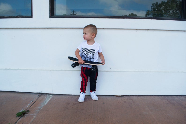 May include: A young boy wearing a white t-shirt with the word "Chaos" printed on it, black and red plaid pants, and white sneakers, holds a wooden skateboard.