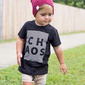 May include: A young child wearing a black t-shirt with white stripes and the word "CHAOS" printed on it. The child is also wearing a burgundy beanie and beige shorts.