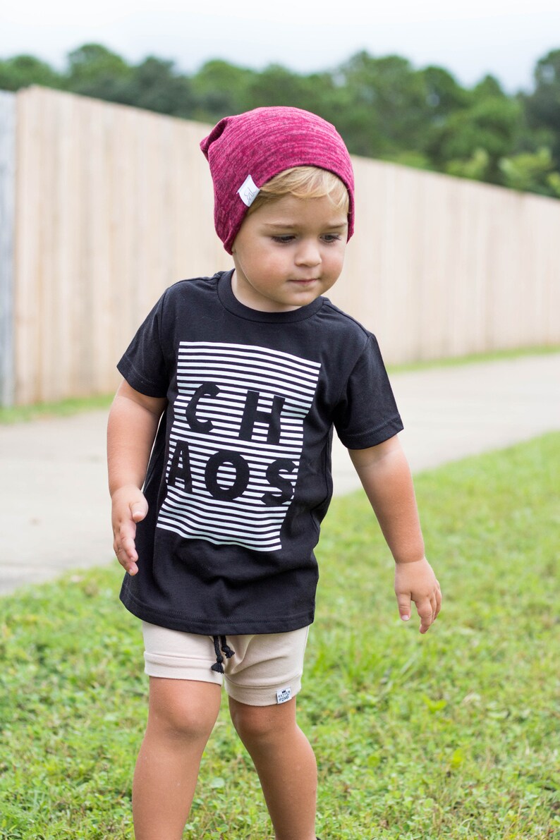 May include: A young child wearing a black t-shirt with white stripes and the word "CHAOS" printed on it. The child is also wearing a burgundy beanie and beige shorts.