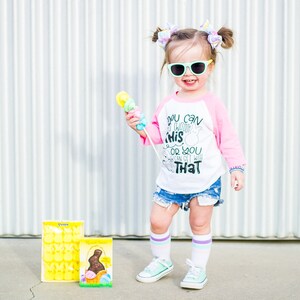 以下が含まれることがあります： サングラスをかけた幼い子供。ピンクと白の野球Tシャツには「YOU CAN THINK THIS OR YOU CAN GET WITH THAT」の文字、デニムショーツ、ミントグリーンのスニーカーを着用。子供はカラフルなおもちゃを持っており、波型の金属壁の前に立っています。