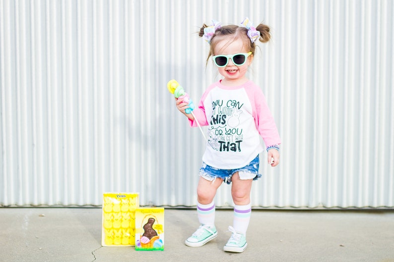 以下が含まれることがあります： サングラスをかけた幼い子供。ピンクと白の野球Tシャツには「YOU CAN THINK THIS OR YOU CAN GET WITH THAT」の文字、デニムショーツ、ミントグリーンのスニーカーを着用。子供はカラフルなおもちゃを持っており、波型の金属壁の前に立っています。