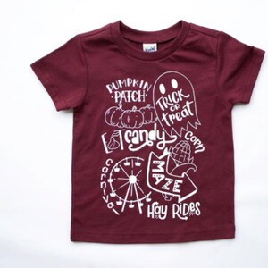 Puede incluir: Camiseta de manga corta color burdeos con un diseño gráfico blanco que representa un huerto de calabazas, maíz dulce, un fantasma, una noria, un laberinto de maíz y el texto "Pumpkin Patch", "Trick or Treat", "Candy Corn", "Maze" y "Hay Rides".