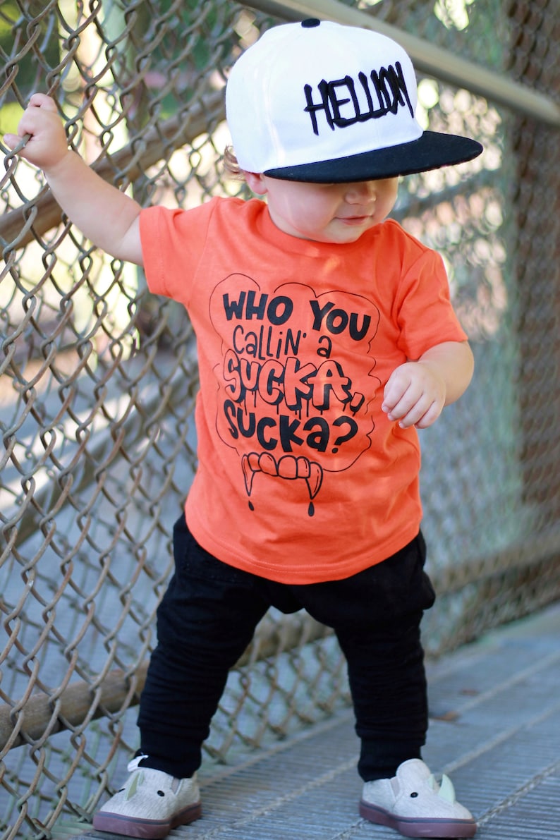 Pu&ograve; includere: Un bambino piccolo che indossa una maglietta arancione con il testo "Who You Callin' a Sucka Sucka?" in nero. Il bambino indossa anche un berretto da baseball nero e bianco con la parola "Hellion" scritta in bianco. Il bambino &egrave; in piedi su una superficie di legno con una recinzione a maglie in background.