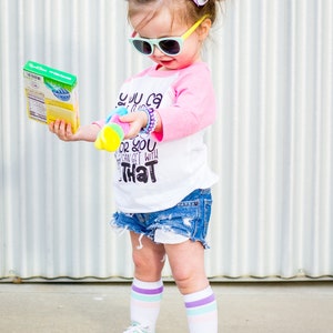 以下が含まれることがあります： サングラスをかけ、「YOU CAN GET WITH THAT」の文字が入った白とピンクのベースボールTシャツ、デニムショーツ、ミントグリーンのスニーカーを履いた幼い子供。子供は箱と玩具を持っています。