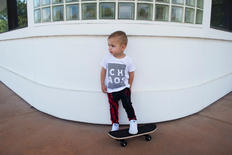 May include: A young child wearing a white t-shirt with black stripes and the text "CH BOSS" printed on it. They are wearing red and black plaid pants and white sneakers. The child is standing on a skateboard.