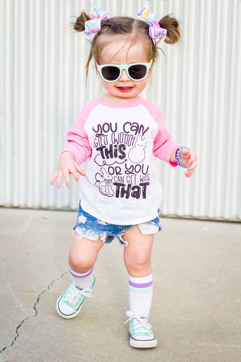 以下が含まれることがあります： サングラスをかけた幼い子供。白とピンクのラグランシャツには「You can get with this or you can get with that」の文字、デニムショーツ、ミントグリーンのスニーカーを着用。子供はストライプの靴下とヘアリボンも身につけています。