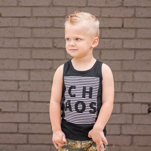 May include: A young boy wearing a black tank top with white stripes and the text "Chaos" printed on it. He is standing in front of a brick wall and wearing camouflage shorts.