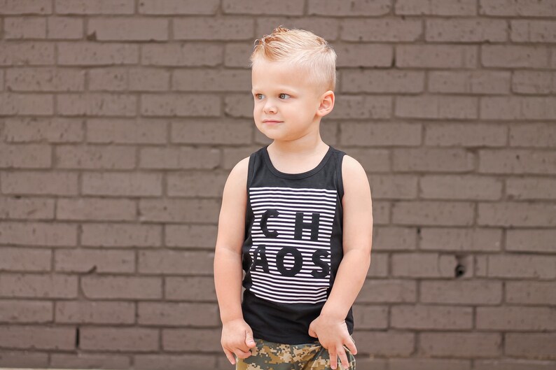 May include: A young boy wearing a black tank top with white stripes and the text "Chaos" printed on it. He is standing in front of a brick wall and wearing camouflage shorts.