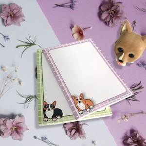 May include: Two pastel pink and green memo pads with a grid pattern. Each pad features a cartoon corgi dog illustration. The text "www.the-pet-patch.com" is printed on the bottom right corner of the pad.