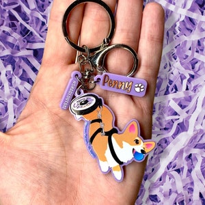 May include: Purple and white keychain featuring a cartoon corgi wearing a harness and pulling a purple tire. The keychain also includes a purple tag with the text "Penny" and a paw print.