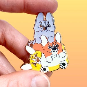 May include: A stack of three enamel pins featuring cartoon dogs. The top dog is light blue with a sparkly finish. The middle dog is orange and white. The bottom dog is yellow and white.
