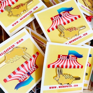 Op de afbeelding: Een set van vier emaillepins met een cartoon corgi-hond die is verkleed als hotdog. De pins zijn geel en goudkleurig met rood-wit gestreepte luifels en blauwe vlaggen met de tekst "Corg Dogs". De pins bevinden zich op een gele achtergrond met de website "www.momopatch.com" die op elke kaart is gedrukt.