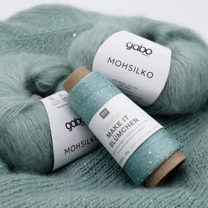 以下が含まれることがあります： 水色の糸とニット生地のクローズアップ。2つの糸玉とスプールには、Gaboの「Mohsilko」とラベルが貼られています。スプールには「creative MAKE IT BLÜMCHEN」とラベルが貼られています。ニット生地はスパンコール付きの水色です。