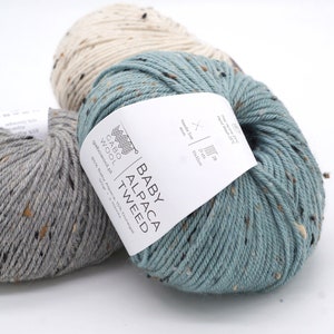 以下が含まれることがあります： グレー、ティール、クリーム色のベビーアルパカツイード糸の3つのかせ。各かせには、色が散りばめられています。ティールのかせには、「BABY ALPACA TWEED」の文字が入った白いラベルが付いています。ラベルには、針のサイズと洗濯方法も記載されています。