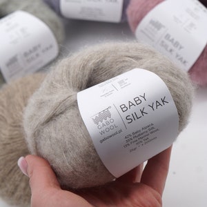 以下が含まれることがあります： ベージュのベビーシルクヤク糸の糸巻き。白いラベルには「ベビーシルクヤク」と糸の構成が記載されています。42% ベビーアルパカ、35% 桑蚕糸、13% ファインメリノウール、10% ヤク。ラベルには「25gr - 200mts」と「gabowool.pl」も記載されています。