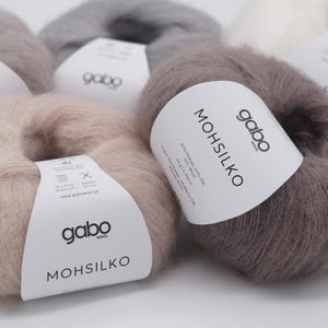 以下が含まれることがあります： ベージュ、グレー、白など、さまざまなニュートラルカラーのGabo Mohsilkoウール糸の複数のかせのクローズアップ。各かせには、ブランド名と製品の詳細が記載された白いラベルが付いています。糸は柔らかくふわふわに見えます。