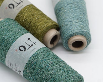 May include: Three conical spools of yarn in shades of green, olive, and teal, each wrapped with a paper band labeled ITO. The yarn features a textured, speckled appearance with small flecks of contrasting color throughout the thread.