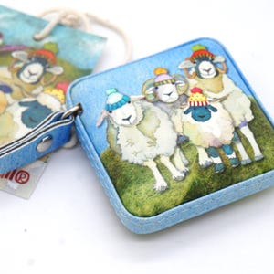 Op de afbeelding: Een vierkante, lichtblauwe portemonnee met een grillige illustratie van schapen met kleurrijke gebreide mutsen. De portemonnee heeft een bijpassende blauwe leren riem en een label met een vergelijkbaar schapenontwerp. De schapen worden afgebeeld op een groen grasveld.