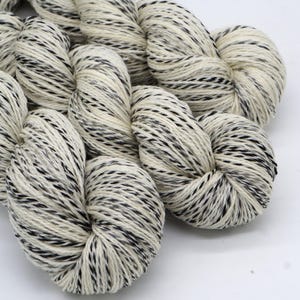 Peut inclure: Gros plan sur plusieurs écheveaux de fil. Le fil est d'un blanc crème avec des mouchetures noires. Le fil est bien enroulé et semble doux et texturé. Le fond est blanc uni.