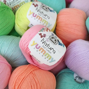 K&ouml;nnte beinhalten: Eine Sammlung bunter Wollkn&auml;uel in den Farben Gr&uuml;n, Gelb, Rosa, Orange und Lila. Jede Kn&auml;uel hat ein Etikett mit der Aufschrift "Natura Just Cotton Yummy Colors" und "Made in EU".