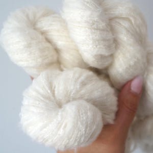 May include: Close-up of a skein of off-white yarn. The yarn is thick and fluffy, with a soft, textured appearance. The yarn is held in a hand, showcasing its natural, undyed colour. Ideal for knitting and crochet projects.