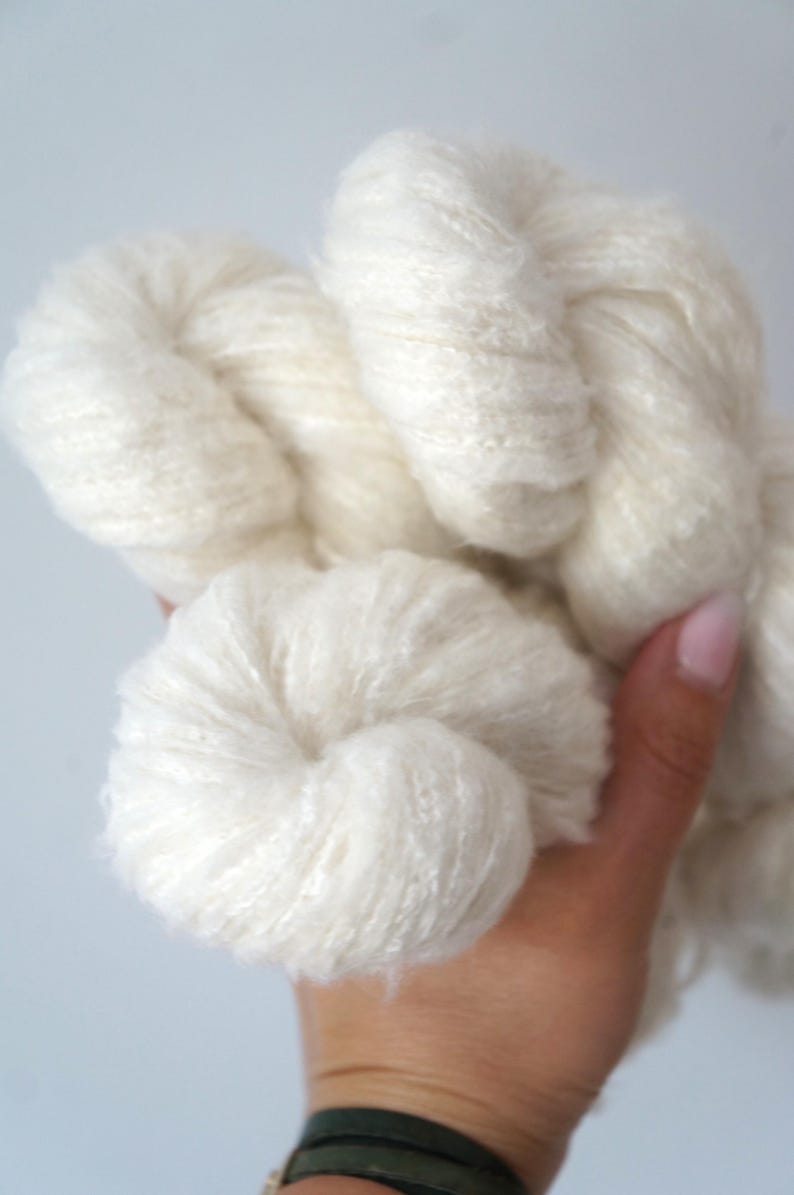 May include: Close-up of a skein of off-white yarn. The yarn is thick and fluffy, with a soft, textured appearance. The yarn is held in a hand, showcasing its natural, undyed colour. Ideal for knitting and crochet projects.