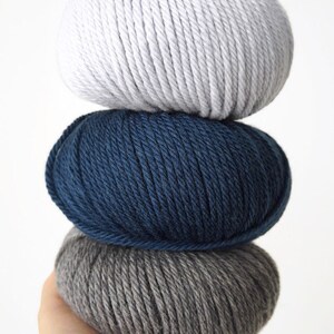 May include: Three balls of yarn stacked on top of each other. The top ball is white, the middle ball is dark blue, and the bottom ball is gray.