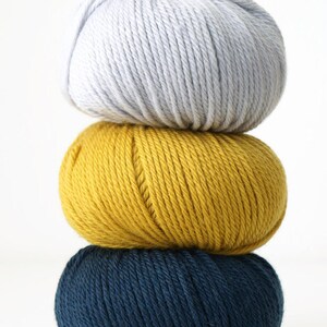 May include: Three balls of yarn stacked on top of each other. The top ball is light gray, the middle ball is yellow, and the bottom ball is dark blue.