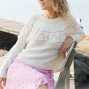 May include: A white knitted sweater with a delicate lace pattern. The sweater has a round neckline and long sleeves. The model is wearing a pink skirt with a floral pattern.