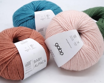 Może przedstawiać: Trzy motki włóczki Gabo Wool Baby Alpaca w kolorach terakoty, jasnego różu i turkusu, z czwartym zielonym motkiem częściowo widocznym. Każdy motek posiada białą papierową etykietę z nazwą marki oraz szczegółami technicznymi.