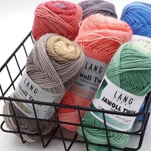 May include: A basket of six skeins of Lang Yarns Jawoll Twin Superwash yarn in various shades of pink, green, blue, and gray. The yarn is labeled with the brand name and the type of yarn.