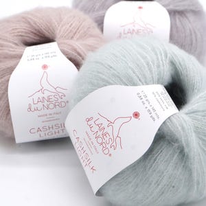 May include: Three skeins of yarn in different colors: light pink, light gray, and light blue. The yarn is labeled "Laines du Nord Cashsilk Light" and is made in Italy. The label also states that the yarn is 70% silk, 22% silk, and 10% cashmere. The skeins are 25 grams each and contain 145 meters of yarn.