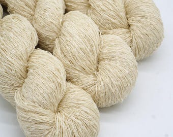 Può includere: Primo piano di diversi gomitoli di filato color naturale. Il filato è di colore beige chiaro e sembra essere fatto di una fibra morbida e strutturata. I gomitoli sono disposti in un mucchio sciolto su uno sfondo bianco, suggerendo un tema artigianale o tessile.
