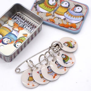 May include: A metal safety pin holding six round stitch markers, each featuring a colourful puffin illustration. The set includes a rectangular metal storage tin with a matching puffin design and the text Emma Ball Stitch Markers printed inside.