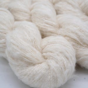 May include: Close-up of off-white yarn, tightly wound into a skein. The yarn has a soft, fluffy texture, with visible fibres. The image is well-lit, highlighting the natural colour and texture of the yarn. Ideal for knitting and crochet projects.