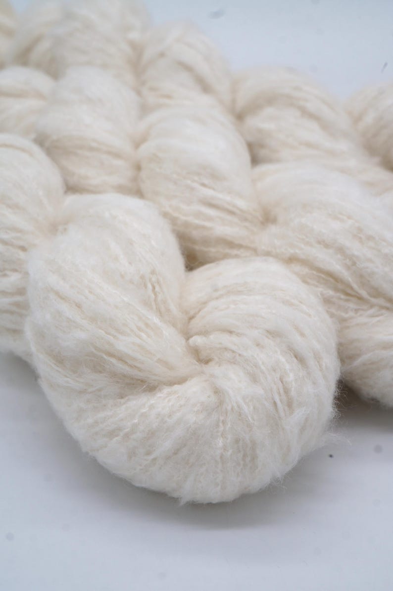 May include: Close-up of off-white yarn, tightly wound into a skein. The yarn has a soft, fluffy texture, with visible fibres. The image is well-lit, highlighting the natural colour and texture of the yarn. Ideal for knitting and crochet projects.