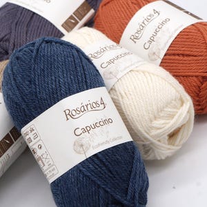 May include: Four balls of yarn in different colours: dark blue, white, orange, and grey. Each ball has a label that reads "Ros&aacute;rios 4 Capuccino Ecofriendly Collection".