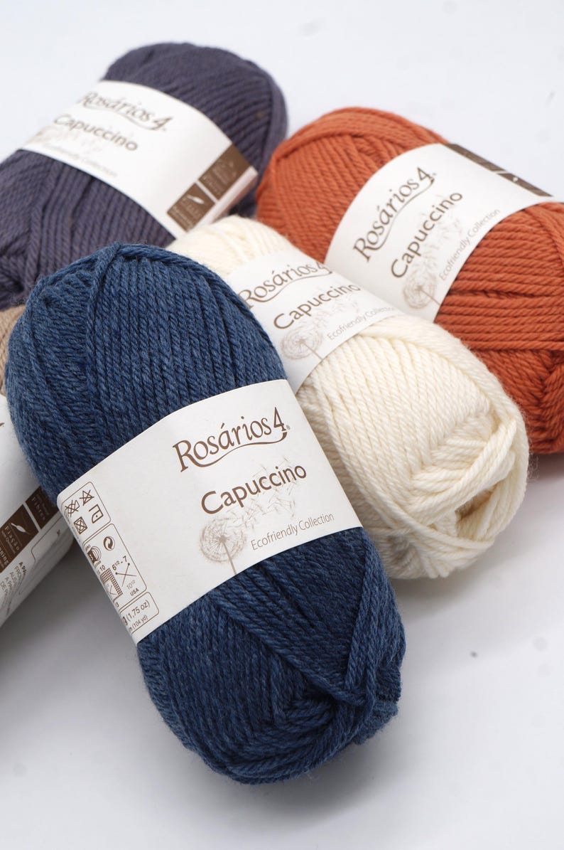 May include: Four balls of yarn in different colours: dark blue, white, orange, and grey. Each ball has a label that reads "Ros&aacute;rios 4 Capuccino Ecofriendly Collection".