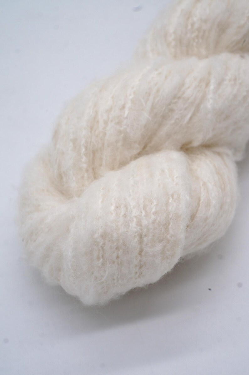 May include: Close-up of a skein of cream-coloured yarn. The yarn has a soft, fluffy texture with a slightly textured surface. The fibres are tightly spun, creating a dense, yet airy appearance. Suitable for knitting or crochet projects.