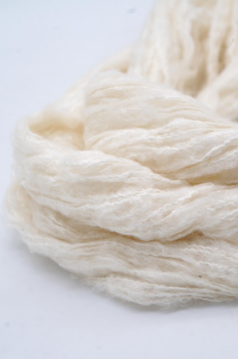 May include: Close-up of a bundle of cream-coloured yarn. The yarn has a soft, fluffy texture and a slightly textured surface. The fibres are loosely spun, giving it a light and airy appearance. Suitable for knitting or crochet projects.