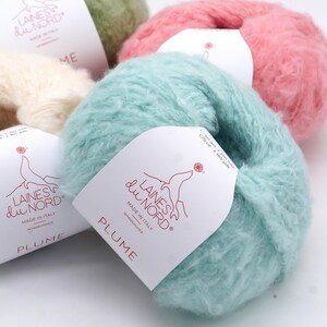 Pu&ograve; includere: Primo piano di diversi gomitoli di filato soffice nei toni del verde, rosa e blu. Ogni gomitolo ha un'etichetta con la scritta "LAINES du NORD" e "PLUME". Prodotto in Italia.