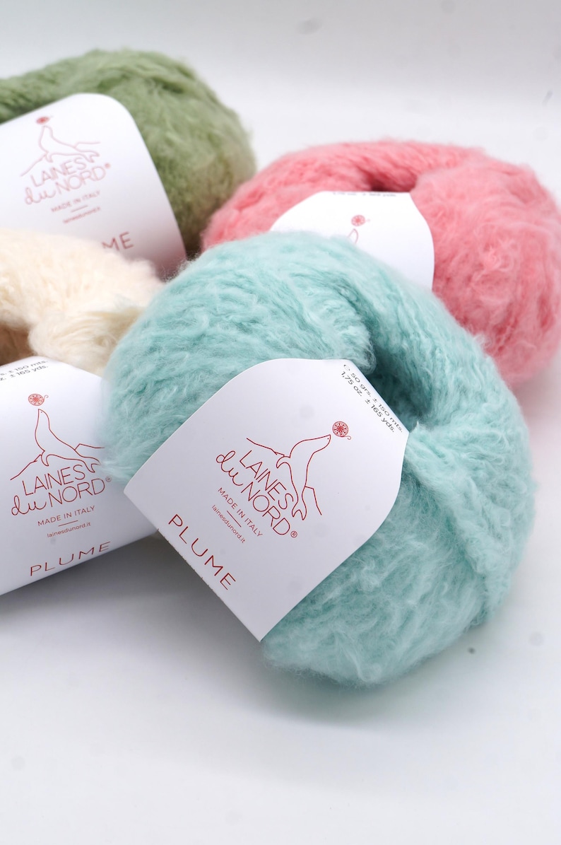 Pu&ograve; includere: Primo piano di diversi gomitoli di filato soffice nei toni del verde, rosa e blu. Ogni gomitolo ha un'etichetta con la scritta "LAINES du NORD" e "PLUME". Prodotto in Italia.