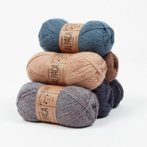 May include: Four skeins of yarn stacked on top of each other. The yarn is a soft, light brown, dark blue, and a mottled blue and gray. The yarn labels are visible and say "DROPS ALPACA UNI COLOUR all-time favorite".
