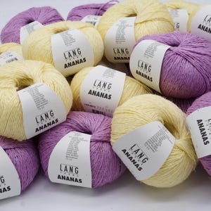 Puede incluir: Un conjunto de madejas de hilo en tonos amarillos y morados. Cada madeja tiene una etiqueta blanca con el texto "LANG ANANAS". El hilo está dispuesto en un montón, con algunas madejas superpuestas.
