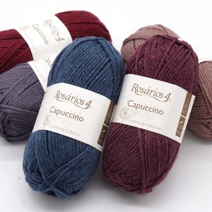 May include: Six balls of yarn in various colours, including burgundy, grey, blue, and brown. Each ball has a label that reads "Ros&aacute;rios 4 Cappuccino" and "Exclusively Collection".