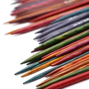 Könnte beinhalten: Eine Sammlung bunter Holzstricknadeln, die fächerförmig angeordnet sind. Die Nadeln bestehen aus verschiedenen Holzarten, jede mit ihrem eigenen einzigartigen Maserungsmuster und ihrer Farbe. Die Nadeln sind spitz und bereit zum Stricken.